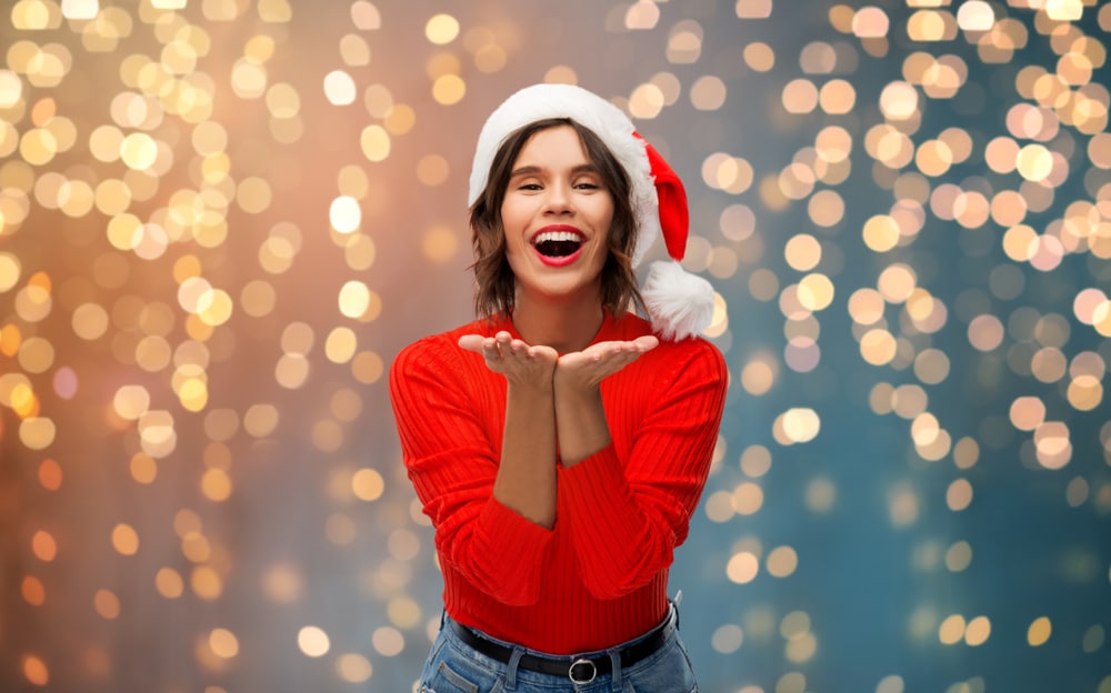 Woman with a beautiful white smile stands in front of twinkling holiday light display wearing a Santa hat